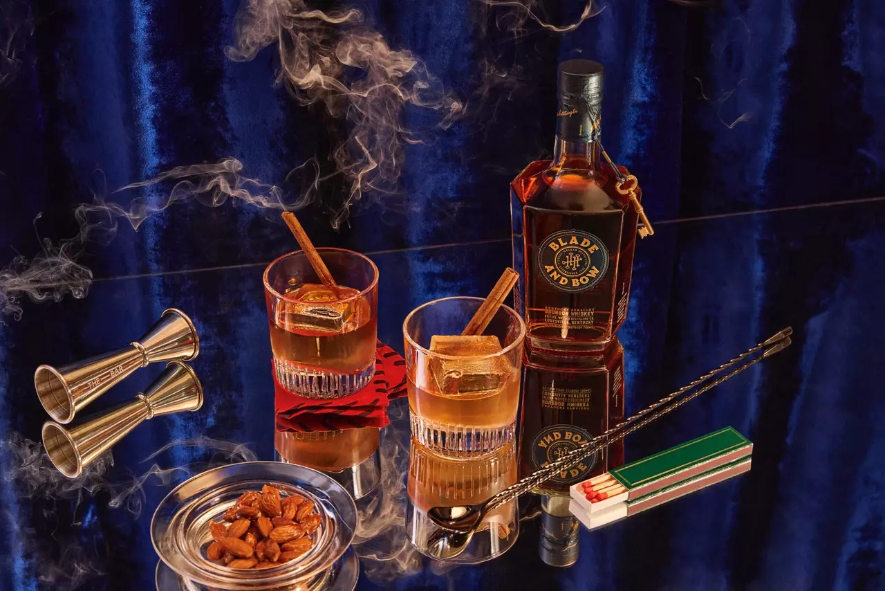 A bottle of Blade and Bow bourbon, two glasses with drinks and cinnamon sticks, a jigger, an ashtray with almonds, matches, a bar spoon, and smoke on a reflective surface.