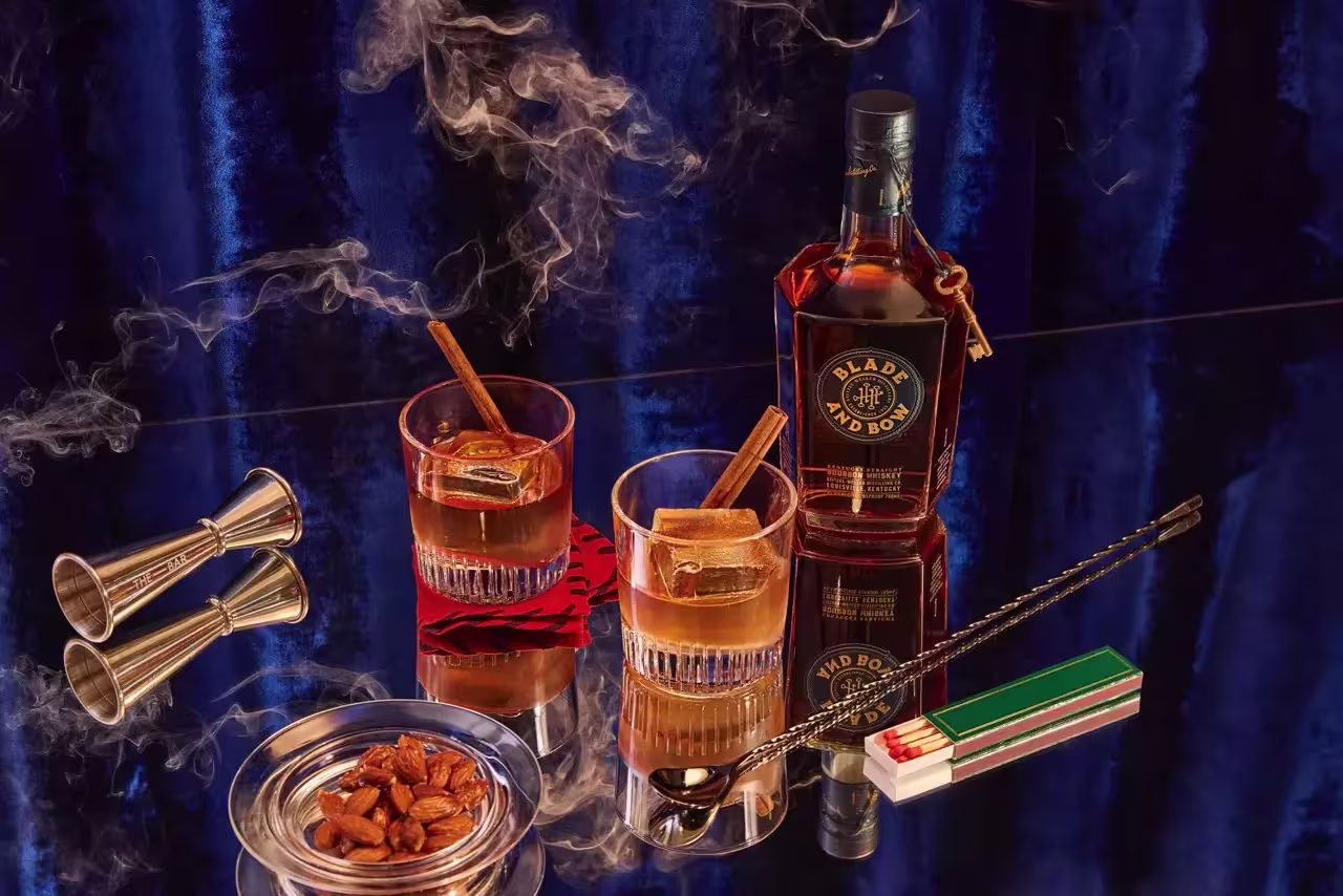 A bottle of Blade and Bow bourbon, two glasses with drinks and cinnamon sticks, a jigger, an ashtray with almonds, matches, a bar spoon, and smoke on a reflective surface.