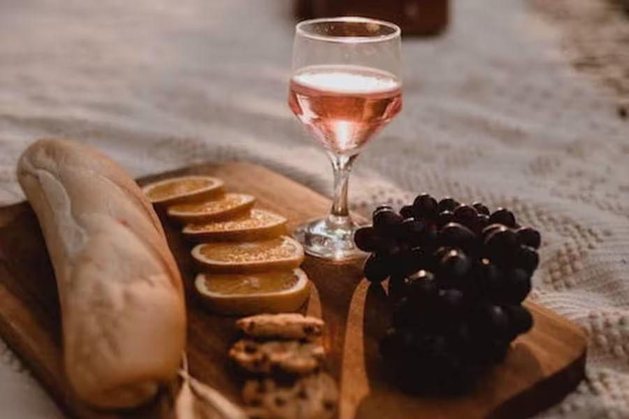 A wooden board with a baguette, orange slices, chocolate chip cookies, grapes, and a glass of pink wine on a white textured surface.
