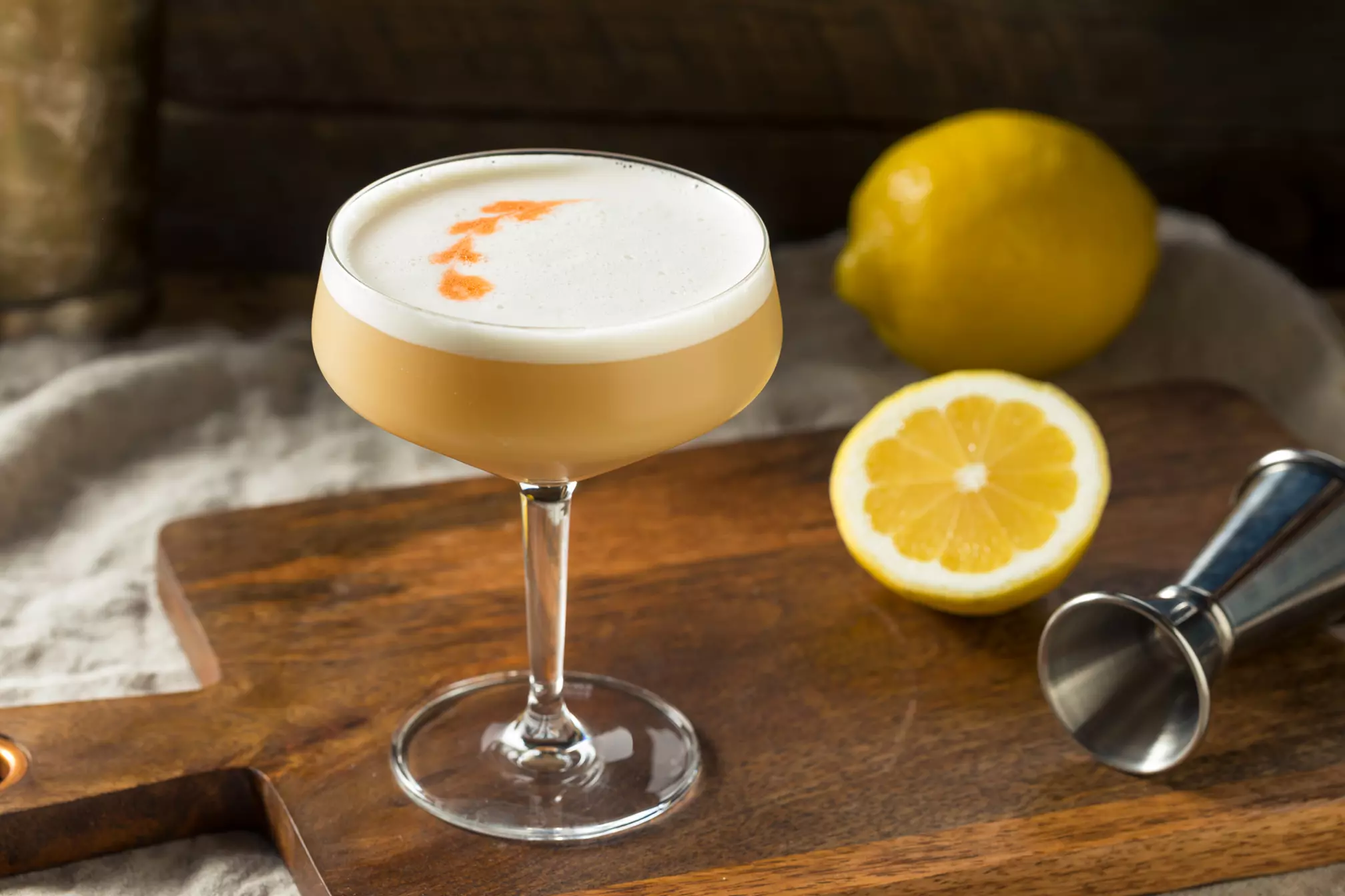 A cocktail with a foamy top and bitters in a coupe glass on a wooden board, next to a halved lemon and a jigger.