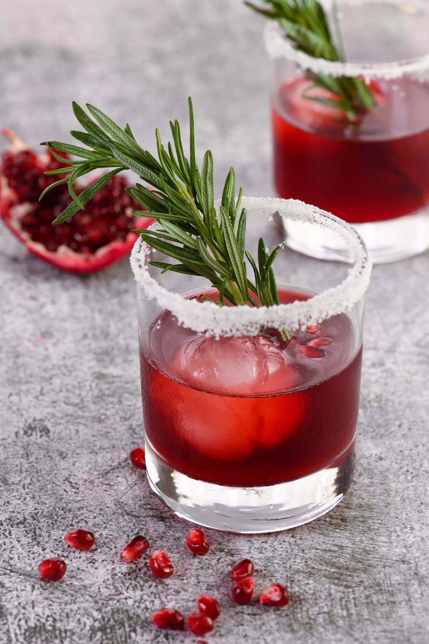 A glass of red cocktail with ice, sugared rim, and a sprig of rosemary garnish; pomegranate seeds and a pomegranate half are visible nearby.