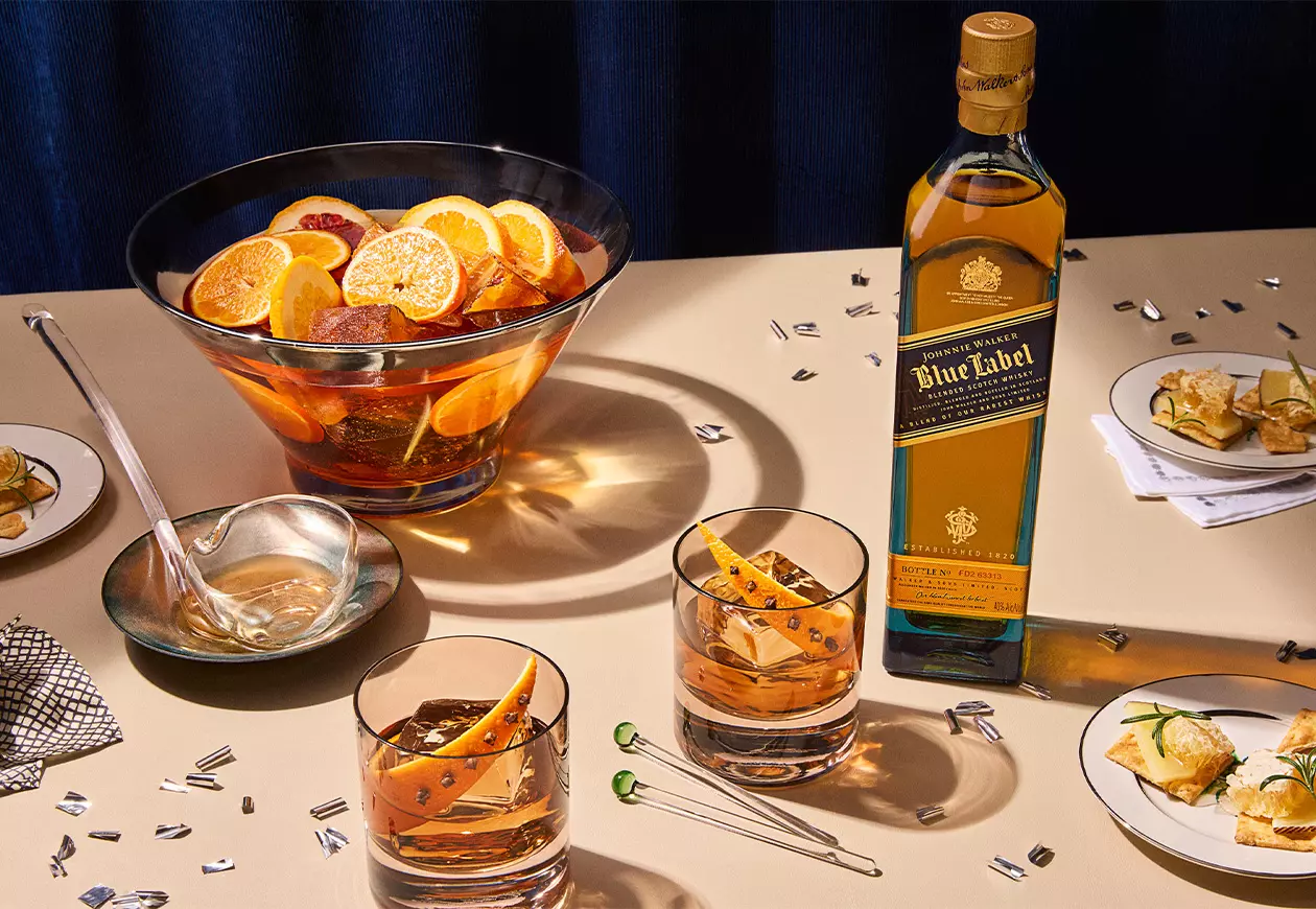 A bottle of Johnnie Walker Blue Label whisky sits on a table with two whisky glasses, a punch bowl with orange slices, and plates of appetizers.