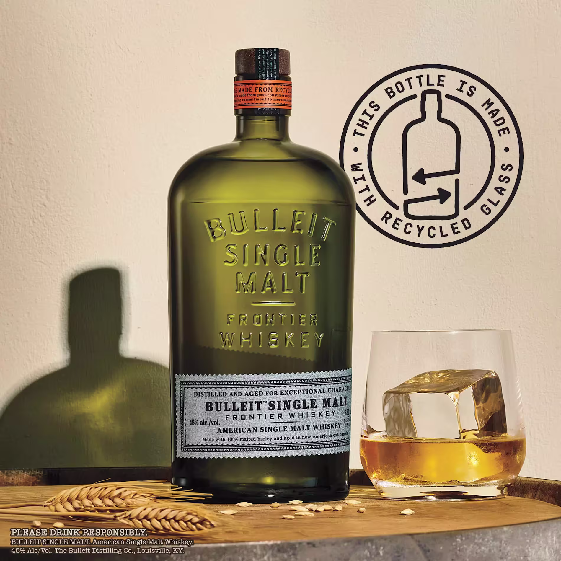 A bottle of Bulleit Single Malt Frontier Whiskey sits on a wooden surface next to a glass of whiskey with ice, with a sign indicating the bottle is made from recycled glass.