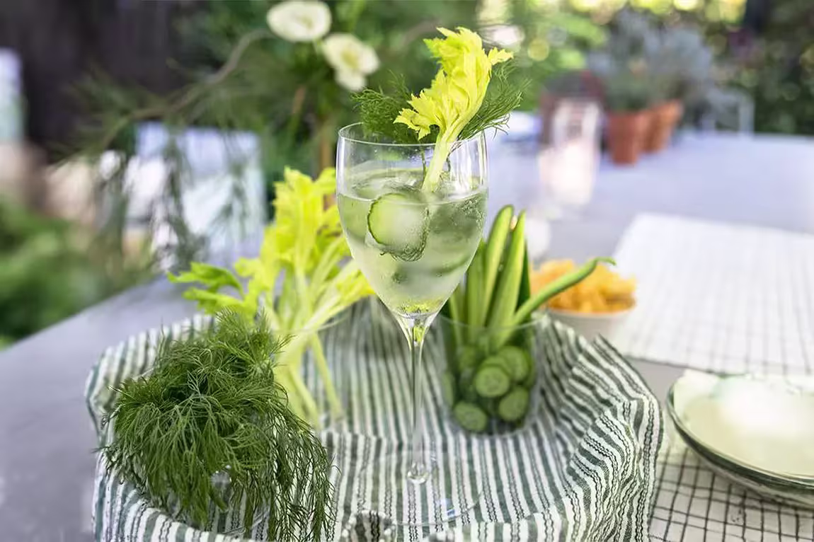 A glass of clear cocktail with ice, garnished with celery, cucumber, and dill, sits on a striped cloth next to fresh herbs and sliced vegetables outdoors.