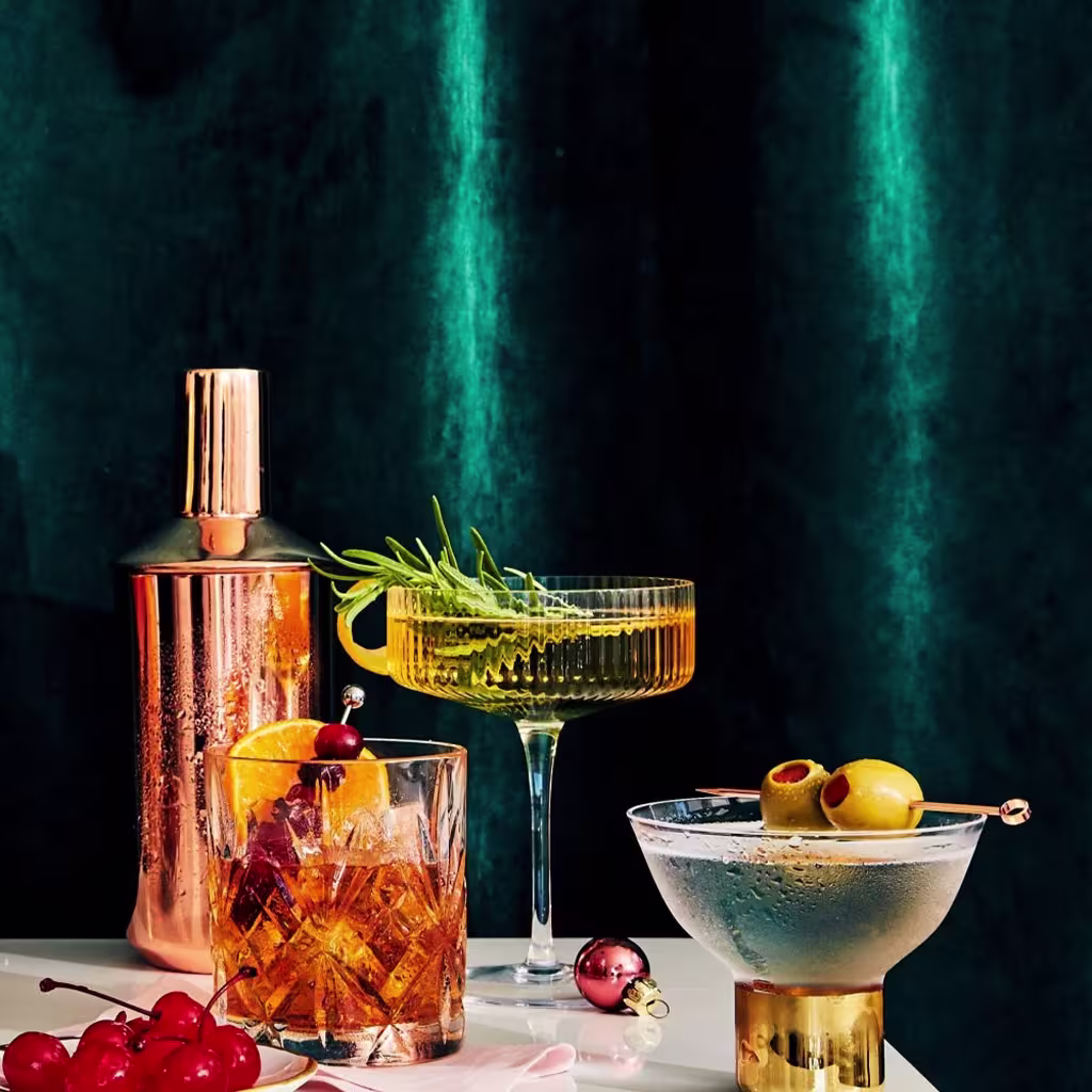 Three cocktails—a whiskey drink with an orange slice, a martini with olives, and a yellow drink with rosemary—sit beside a copper shaker and cherries on a white surface with a dark green backdrop.