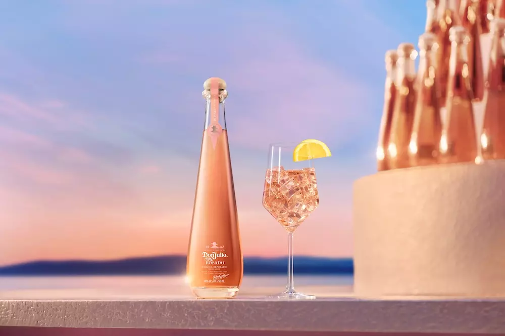 A bottle of Don Julio Rosado tequila and a glass with a lemon wedge sit on a smooth surface, with several bottles and a sunset view in the background.