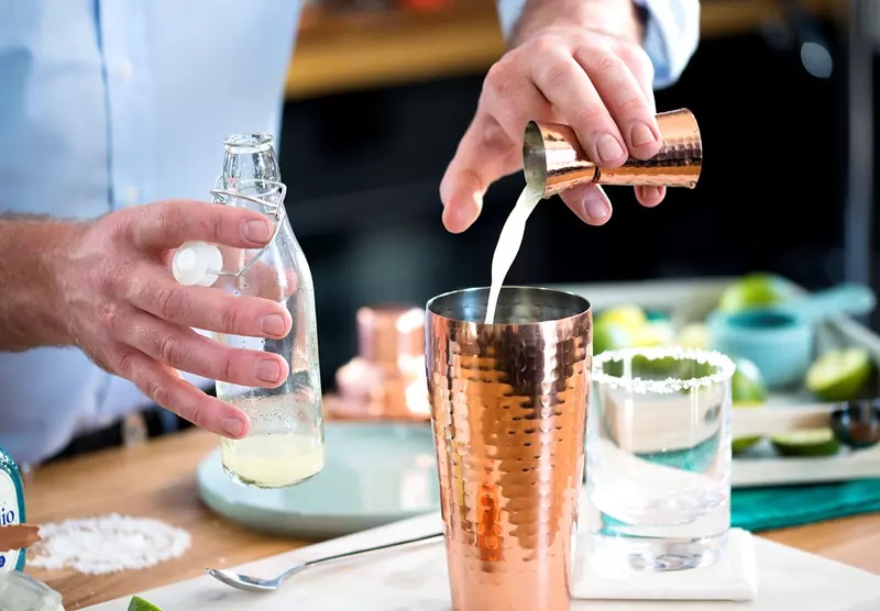 A person pours a white liquid from a copper measuring cup into a cocktail shaker, with a glass bottle, salted-rim glass, and lime slices on the counter.