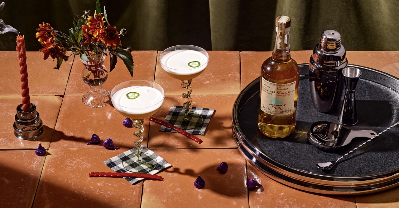 A table with two cocktails garnished with jalapeño slices, a whiskey bottle, cocktail shaker, bar tools, flowers, candles, and scattered wrapped chocolates.