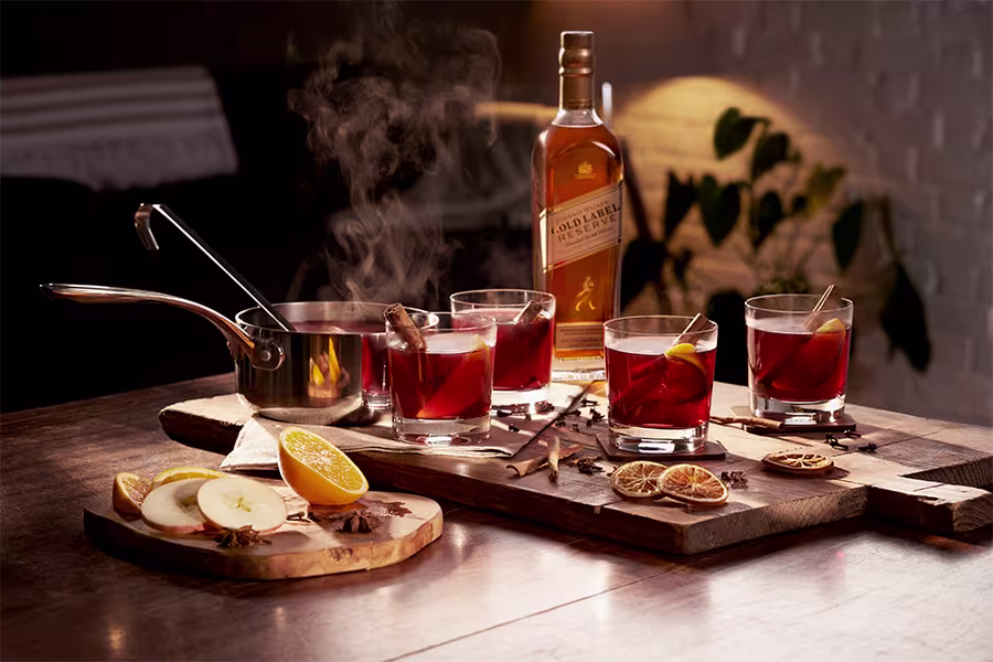 A bottle of whisky, three glasses of hot spiced drink with citrus and cinnamon, a saucepan with steam, and sliced fruit on a wooden table.