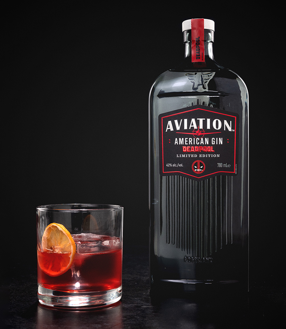 A bottle of Aviation Deadpool gin sits next to a Negroni cocktail in a rocks glass with an orange wheel.