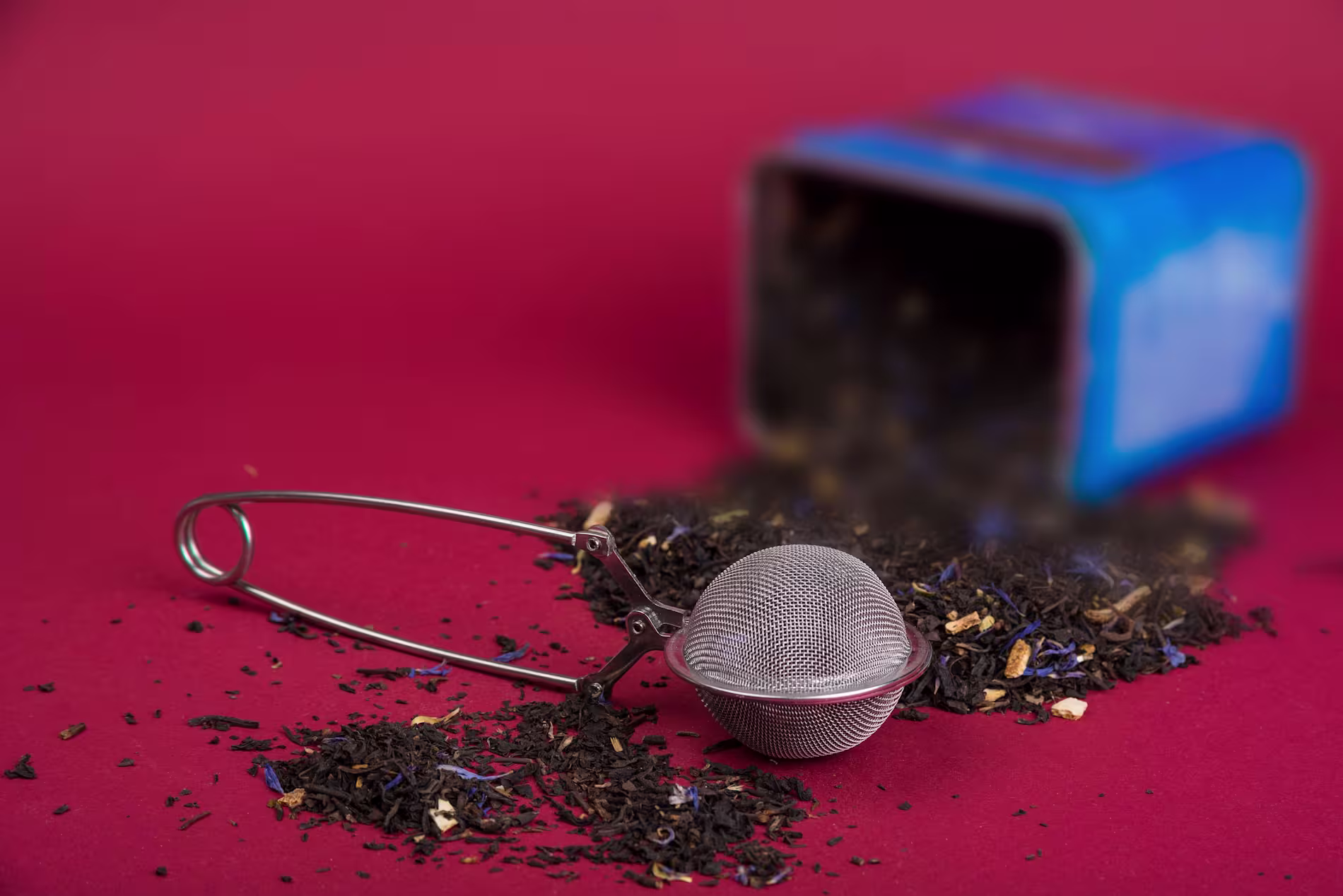 A metal tea infuser rests on loose tea leaves spilled from a blue tin container onto a red surface.
