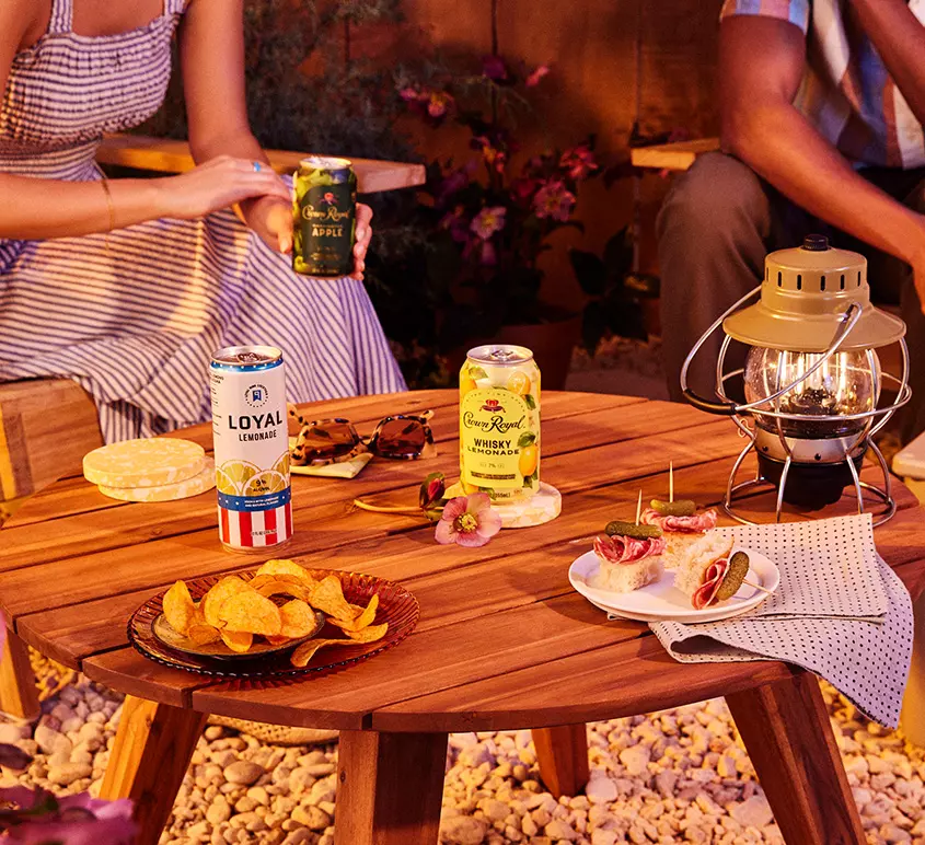 A wooden table outdoors holds canned drinks, snacks, a lantern, and a napkin, with two people seated nearby.