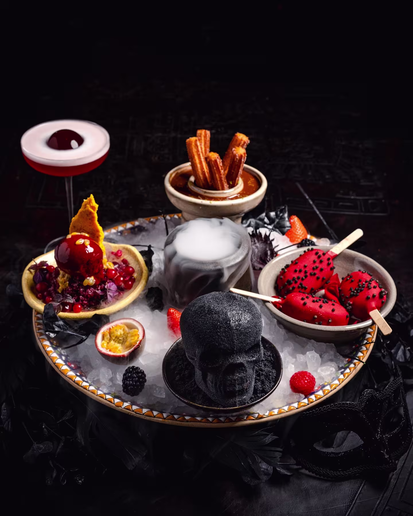 Un surtido de postres artísticos con frutas, churros y una calavera negra, acompañados de una bebida sofisticada en Coya Barcelona.