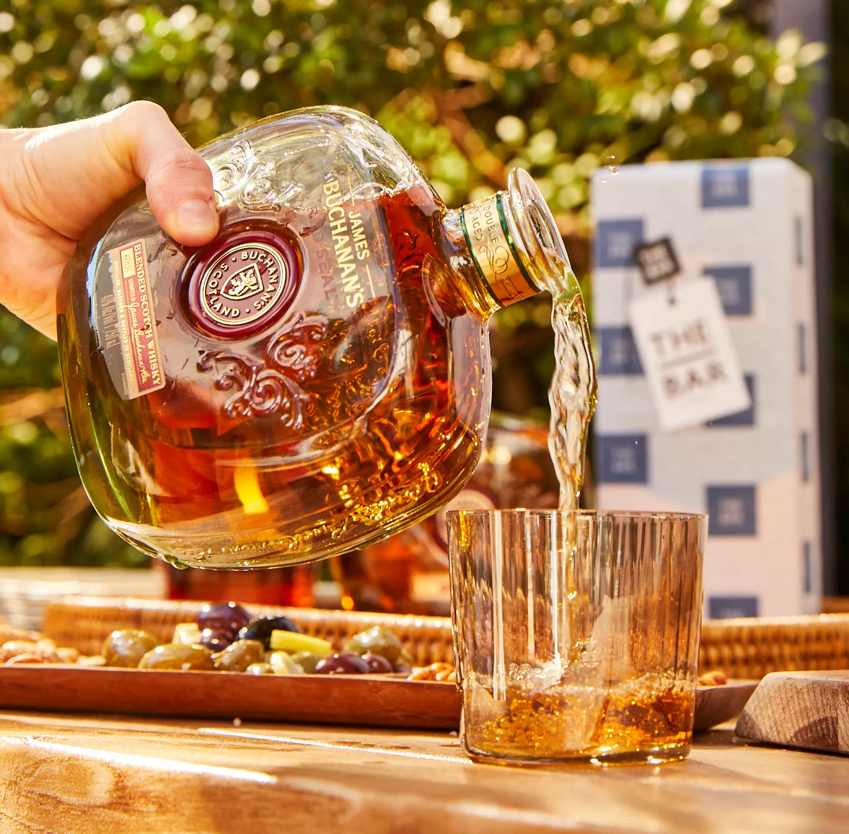 A hand pours whisky from a Buchanans bottle into a glass on a table with snacks. A box and greenery are visible in the background.