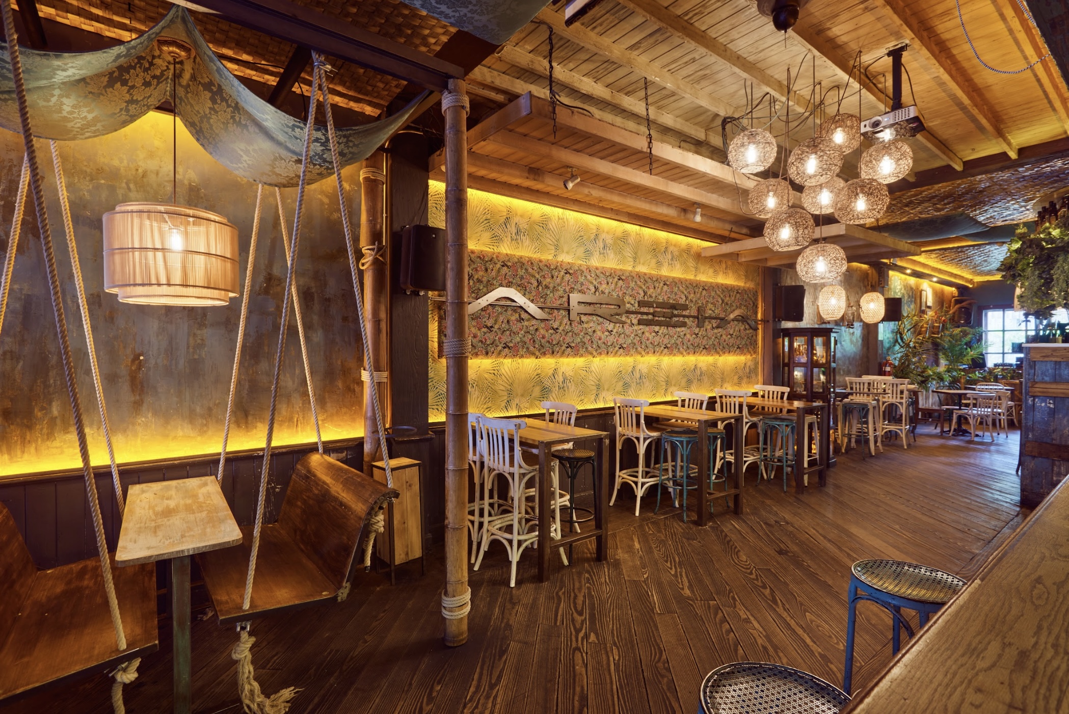 Interior de Areia, con columpios de madera, mesas altas, decoración tropical, luces cálidas y un techo con detalles rústicos y vegetación.