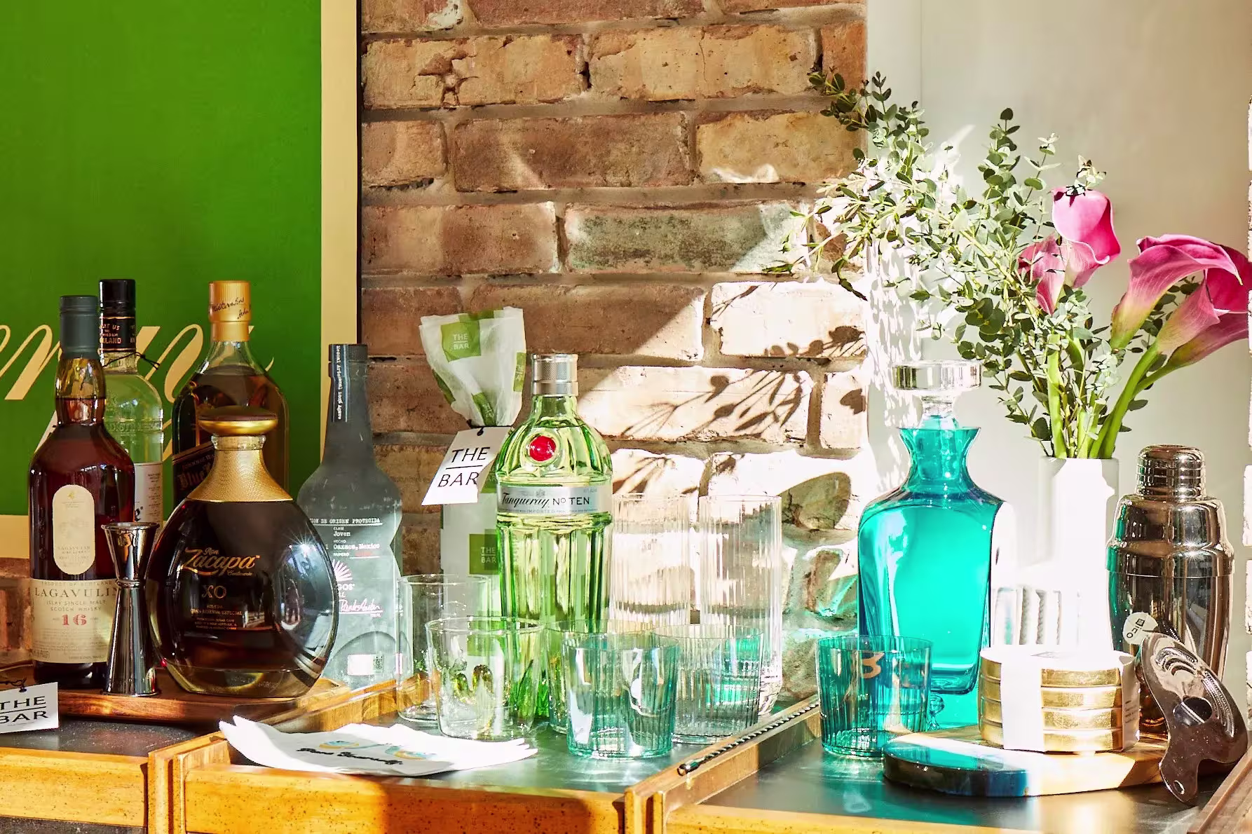 A variety of liquor bottles, glassware, and bar tools arranged on a wooden bar cart beside a brick wall with sunlight and pink flowers.