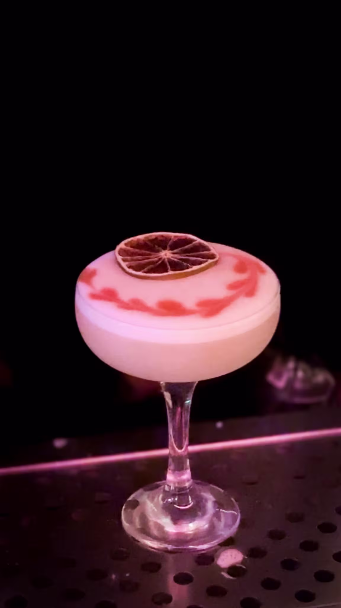 Un cóctel espumoso decorado con un toque de arte rosa y una rodaja de cítrico deshidratado, servido en una copa elegante sobre barra metálica.