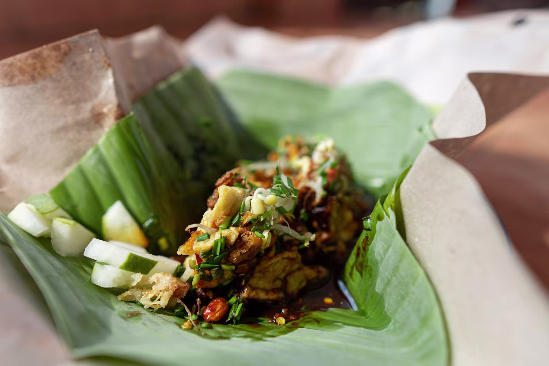 A serving of traditional Indonesian food wrapped in a banana leaf, featuring fried tempeh, sliced cucumber, sprouts, and sauce.