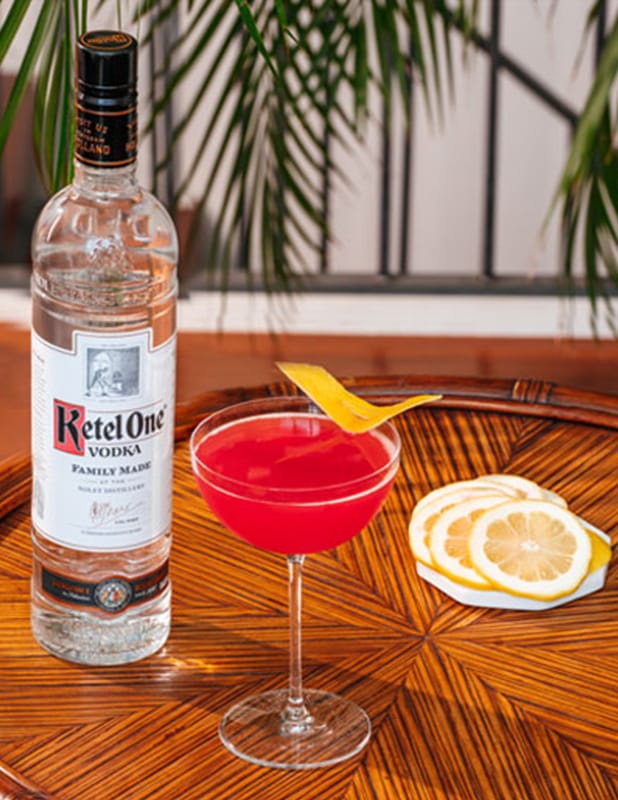 A bottle of Ketel One vodka next to a red cocktail garnished with a lemon twist, with lemon slices on a napkin, on a wooden table.