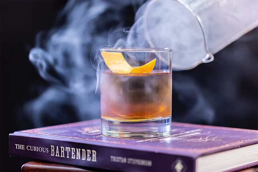 A glass of cocktail with an orange peel garnish sits on a book titled The Curious Bartender, with smoke being poured over the drink from a pitcher.