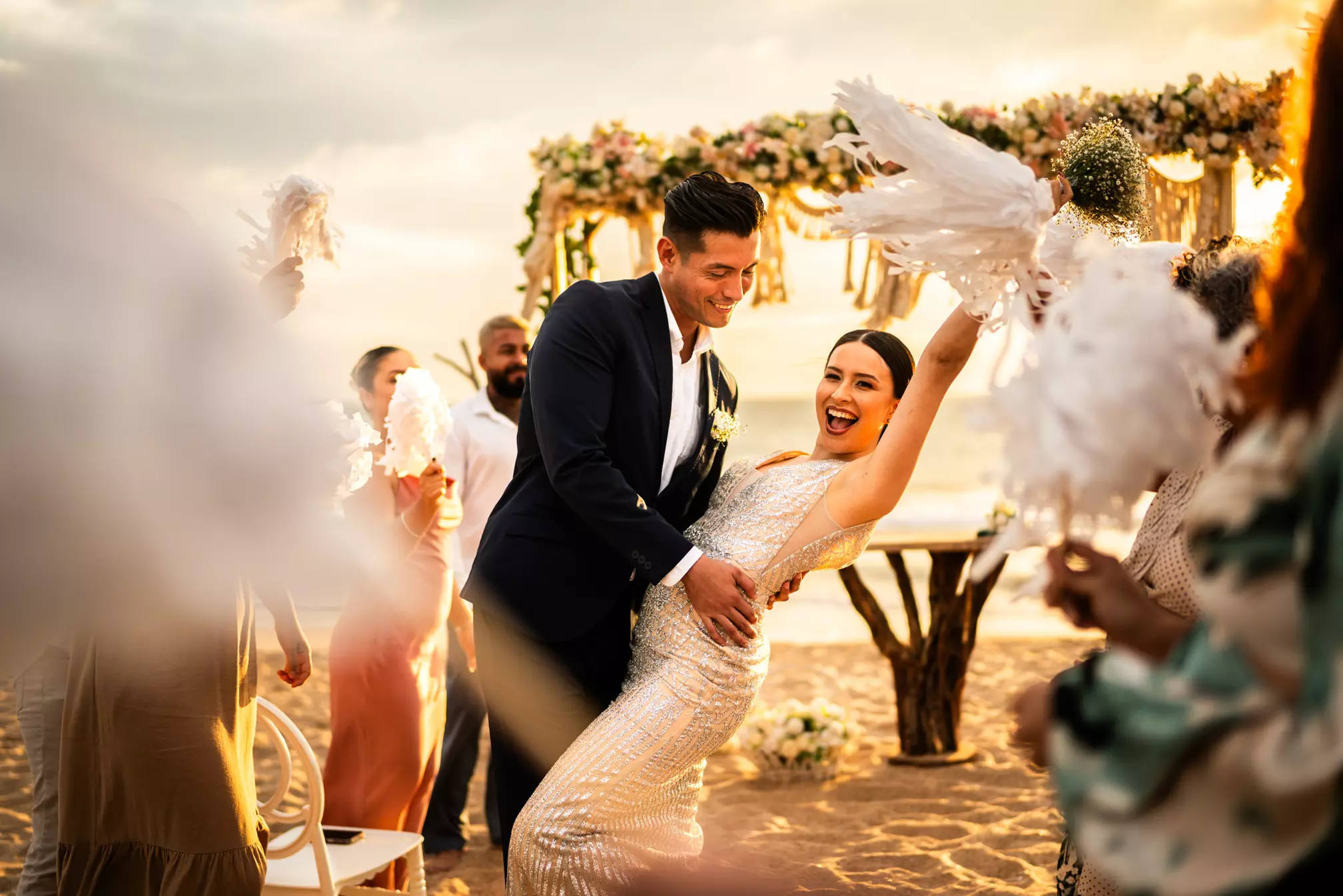 A bride and groom smile and dance together on a beach at sunset, surrounded by guests waving white streamers near a flower-decorated altar.