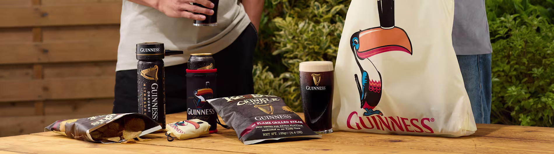 Guinness márkájú termékek egy faasztalon, köztük chipsek, italok és táska.