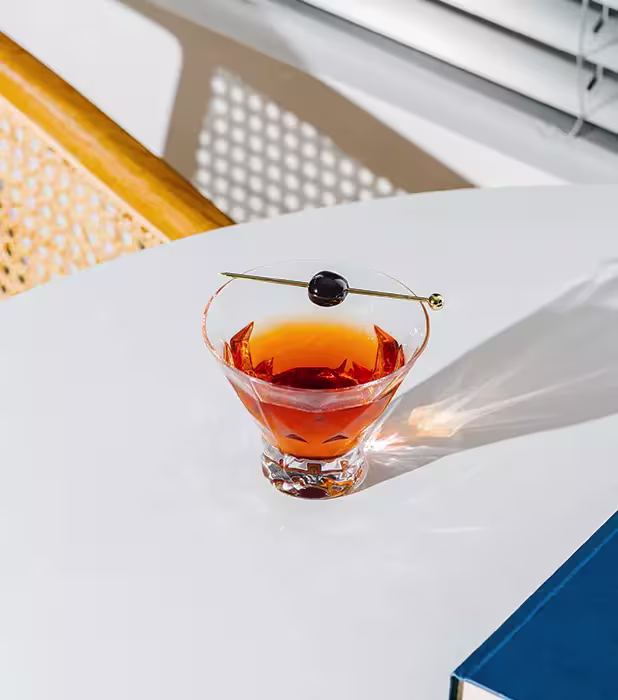 A cocktail with an orange hue, garnished with a dark cherry on a pick, sits on a white table near a book and a woven chair, with sunlight casting shadows.