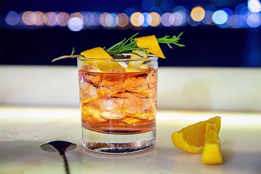 A glass of cocktail with ice, orange peel garnish, and a sprig of rosemary on a bar counter, with orange slices and a spoon nearby, and blurred city lights in the background.