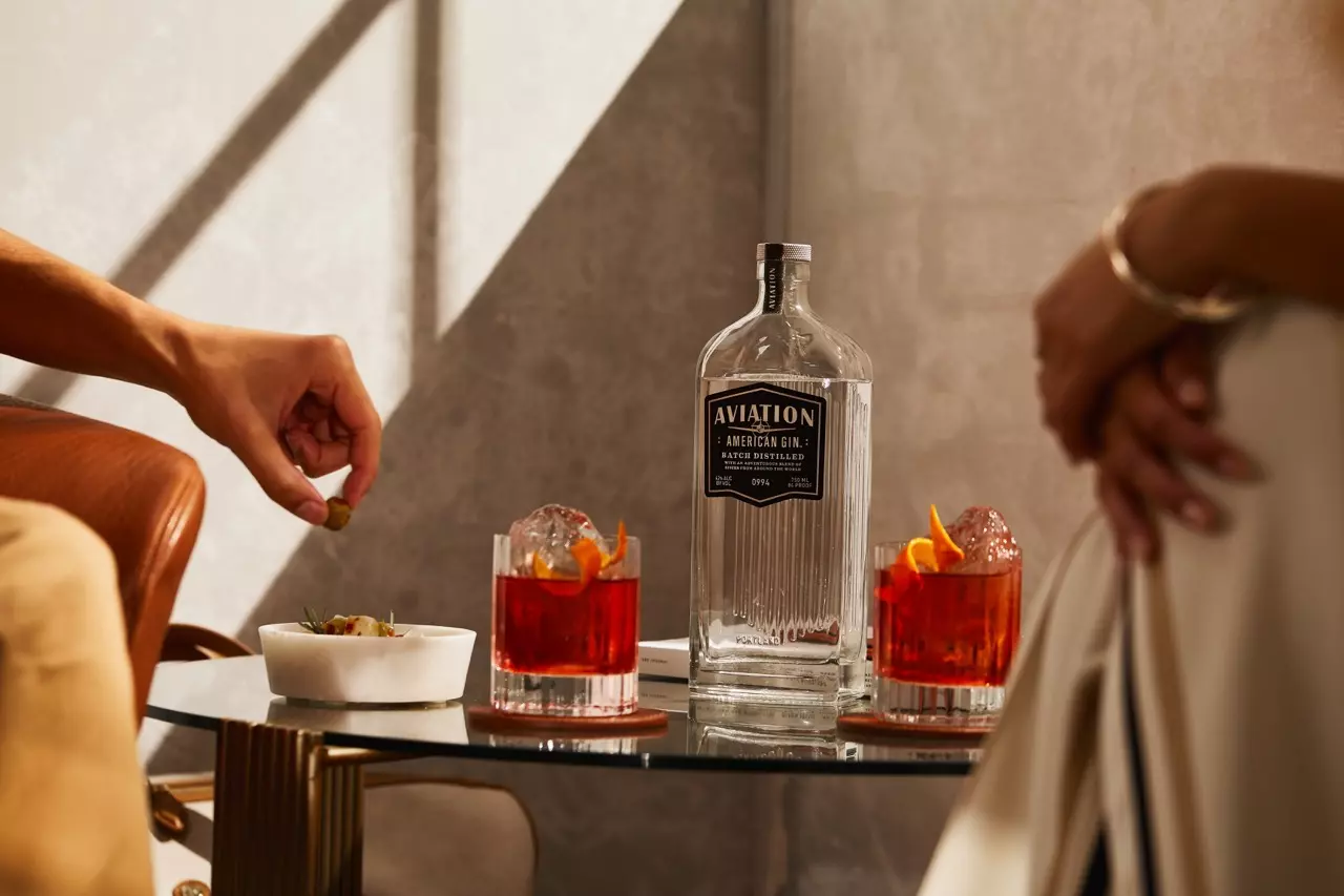 A bottle of Aviation American Gin with two cocktails garnished with orange peels and a bowl of snacks on a glass table, with two people seated nearby.