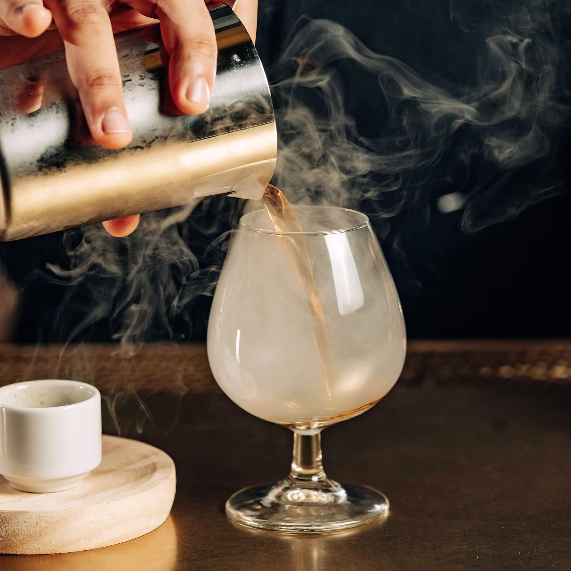 Cóctel servido en copa balón con humo, vertido desde coctelera metálica en un ambiente elegante y oscuro.