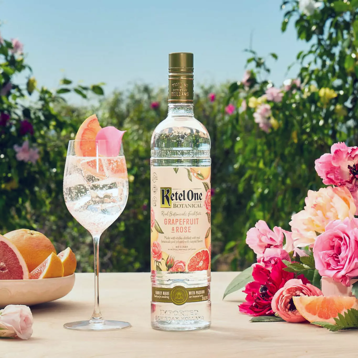 A bottle of Ketel One Botanical Grapefruit & Rose stands on an outdoor table next to a glass cocktail, fresh grapefruit, and pink flowers, with greenery in the background.