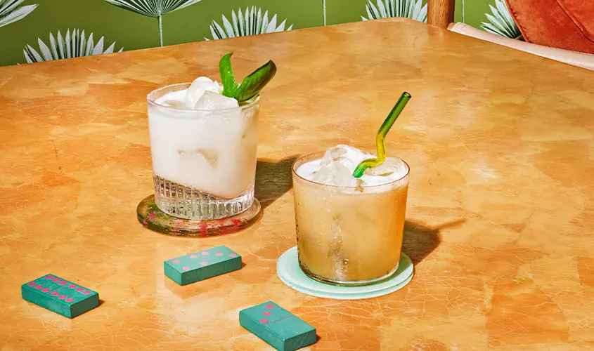 Two cocktails with ice and green garnishes are placed on coasters atop a marble table, with domino tiles nearby and a leafy green wallpaper in the background.