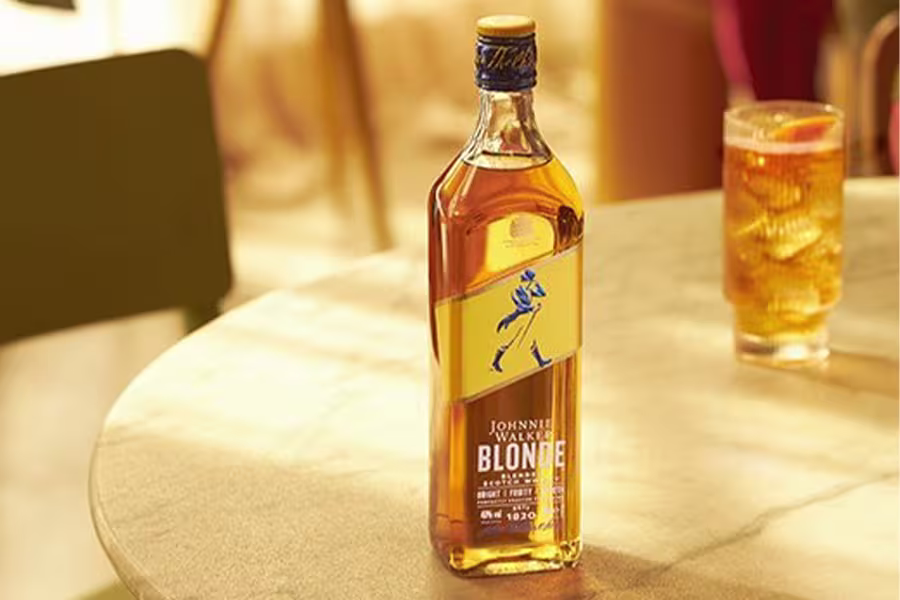 A bottle of Johnnie Walker Blonde whisky is placed on a sunlit table next to a glass filled with an amber-colored drink and ice.