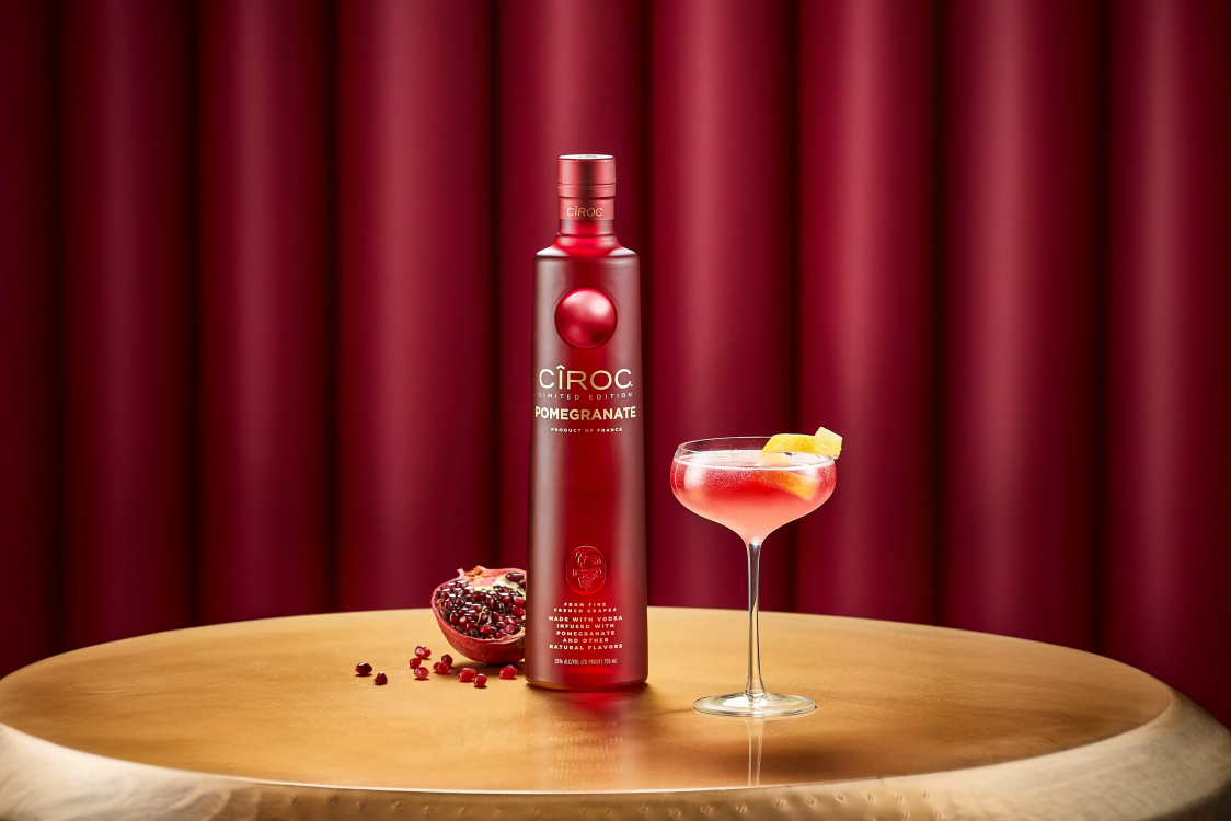 A bottle of Ciroc Pomegranate vodka stands next to a halved pomegranate and a cocktail garnished with lemon on a gold table with a red background.
