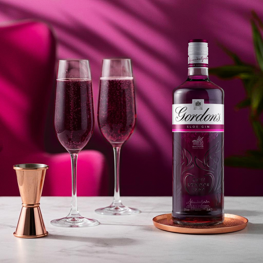 Gordon's Sloe Gin bottle next to two flutes of sloe gin cocktail on a pink background