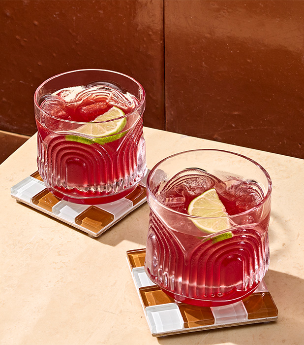 Two glasses of red beverage with ice and lime slices on patterned coasters, placed on a light-colored surface next to a brown wall.