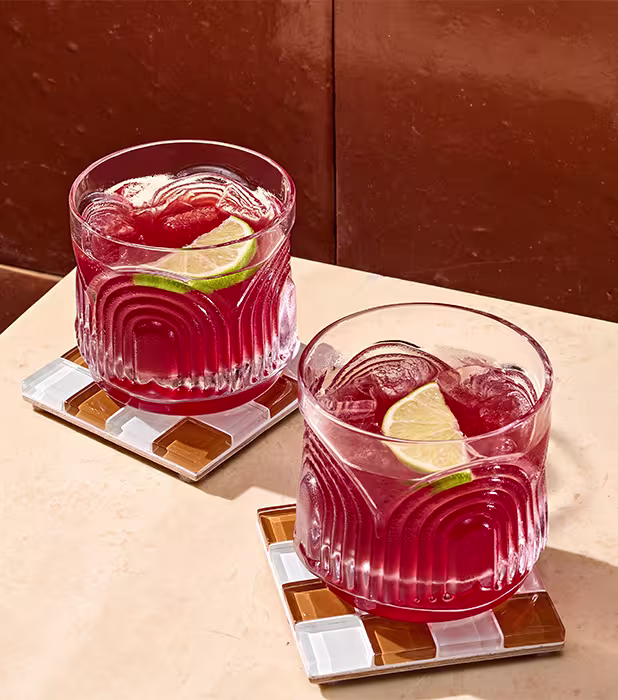 Two glasses of red beverage with ice and lime slices on patterned coasters, placed on a light-colored surface next to a brown wall.