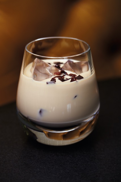 A glass of creamy liqueur with ice cubes and chocolate shavings on top, placed on a dark surface.
