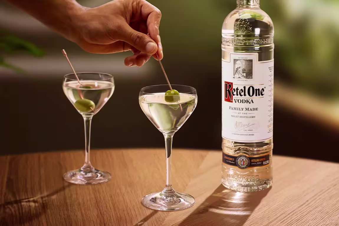 A hand places an olive into a martini glass next to another martini and a bottle of Ketel One vodka on a wooden table.