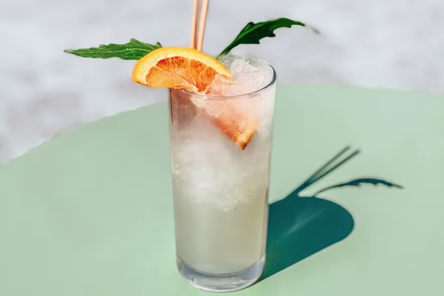 A tall glass filled with a light-colored iced drink, garnished with an orange slice and a leaf, sits on a round green table.