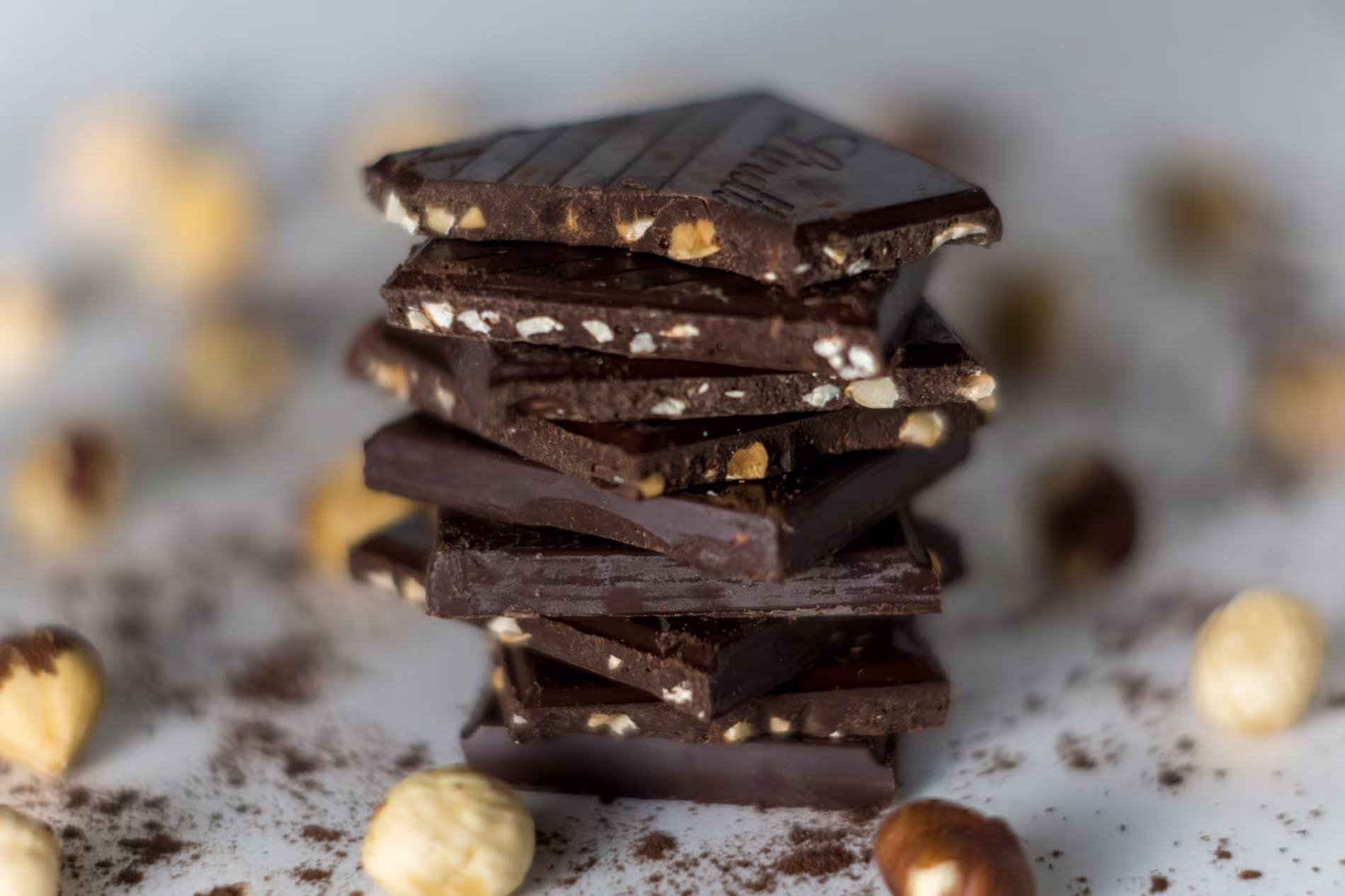 A stack of dark chocolate pieces with hazelnuts, surrounded by scattered whole hazelnuts and cocoa powder on a white surface.