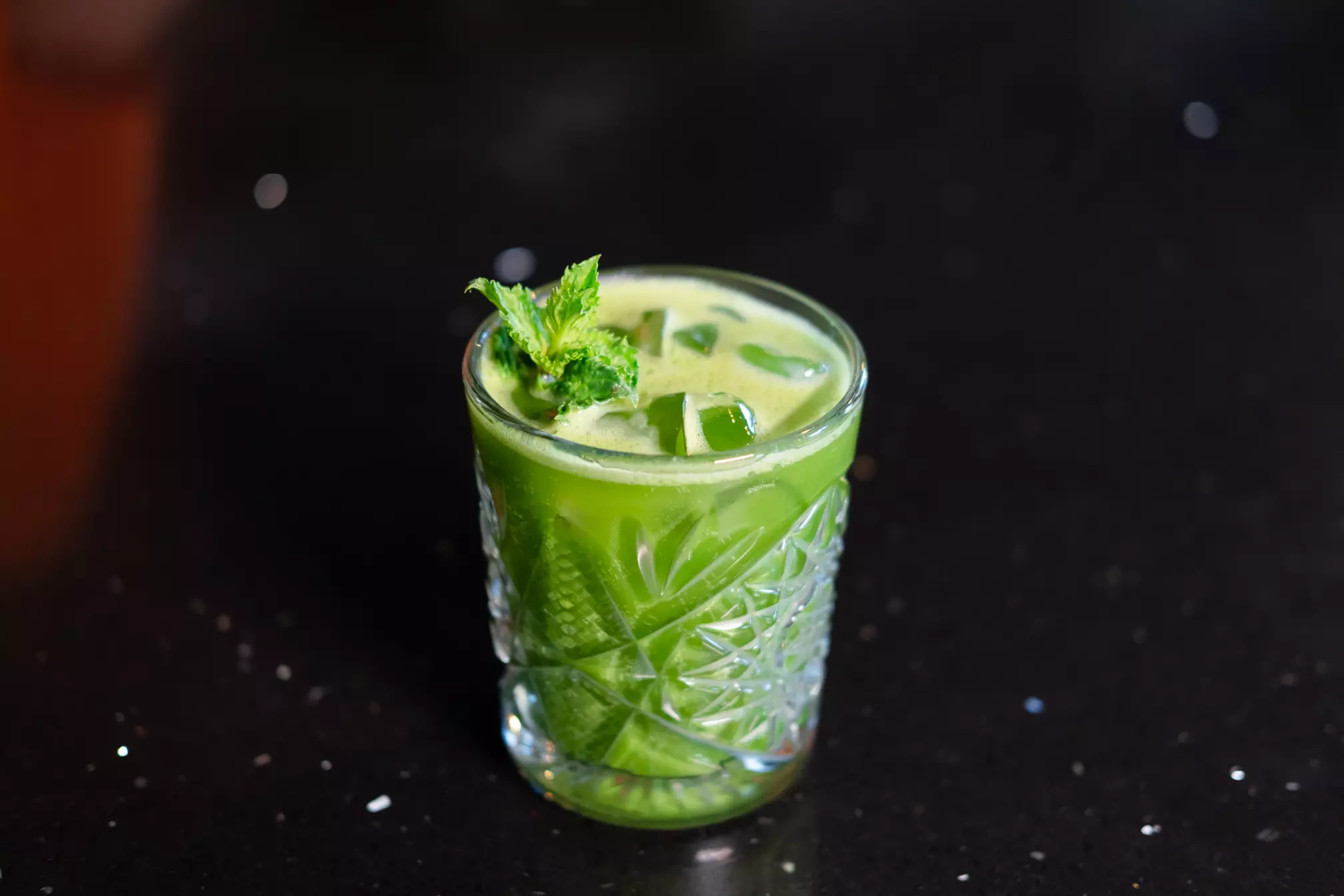 A glass of green juice with ice cubes and a mint garnish sits on a dark surface.