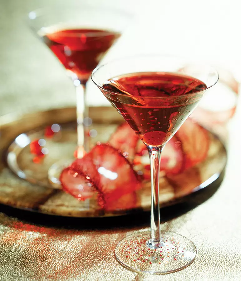 Two martini glasses filled with a red sparkling drink sit on a wooden tray with red-rimmed ice cubes, against a light, shimmering background.