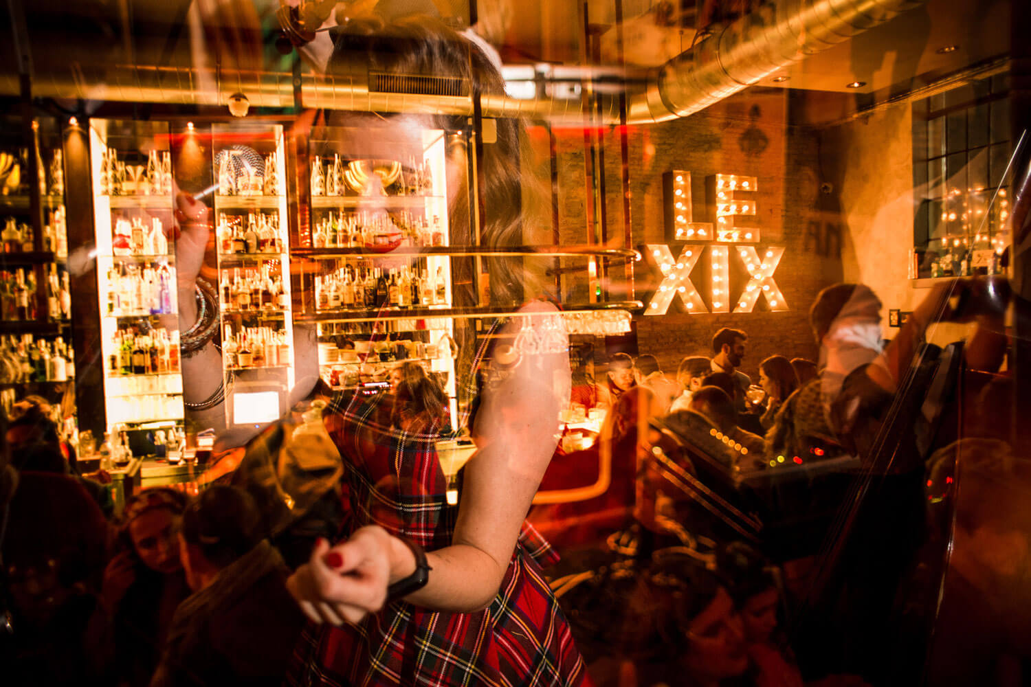 Interior de Le XIX con estanterías llenas de botellas, cartel luminoso en la pared y reflejos de gente bailando y disfrutando del ambiente.