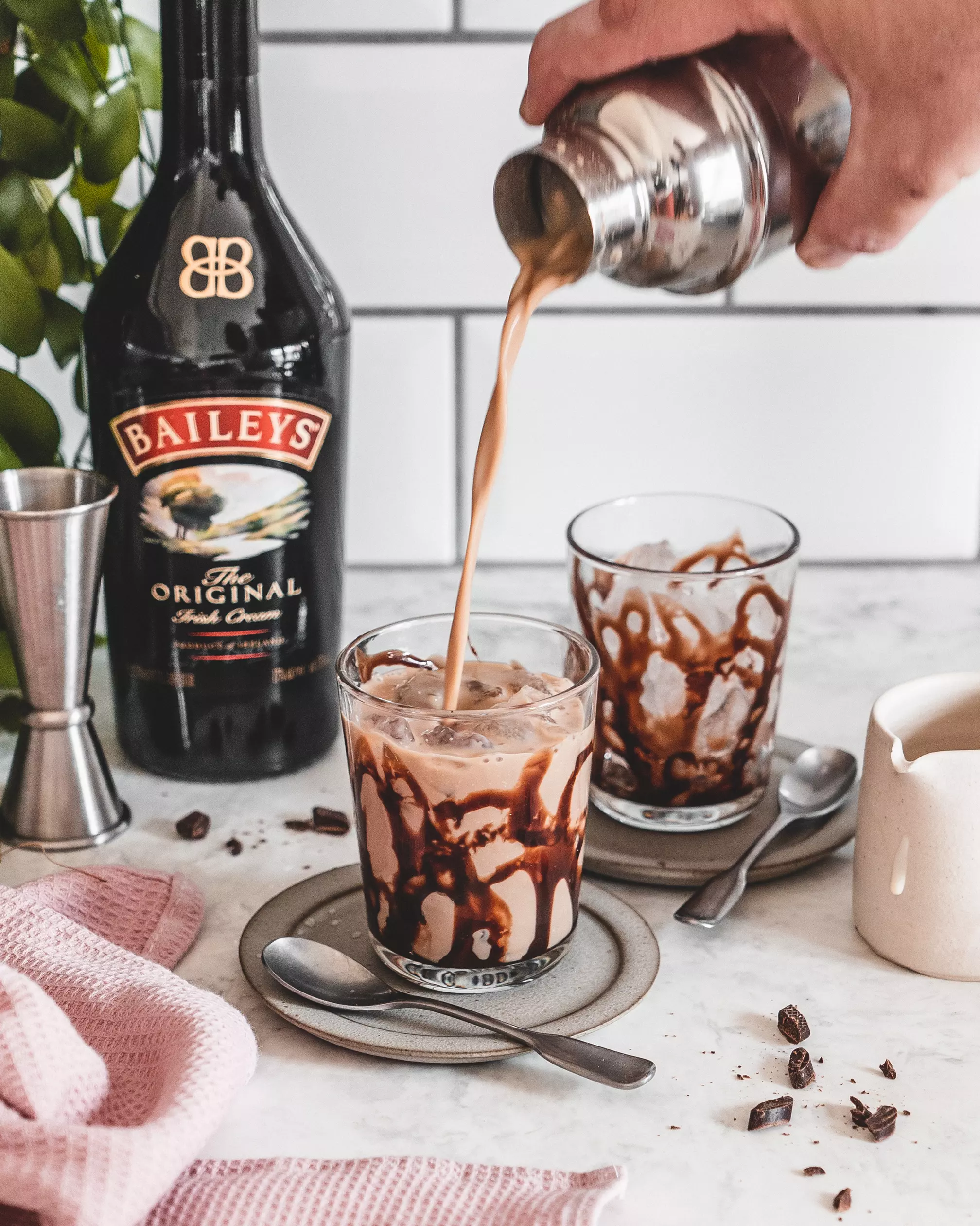 A hand pours Baileys Irish Cream cocktail over ice and chocolate syrup in a glass, with a Baileys bottle, jigger, and napkin nearby on a countertop.