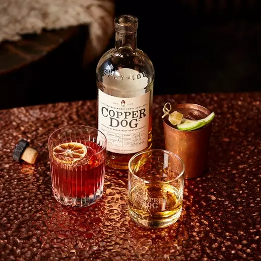 A bottle of Copper Dog whisky on a textured table, with its cap off, surrounded by two whisky glasses and a copper cup with garnishes.