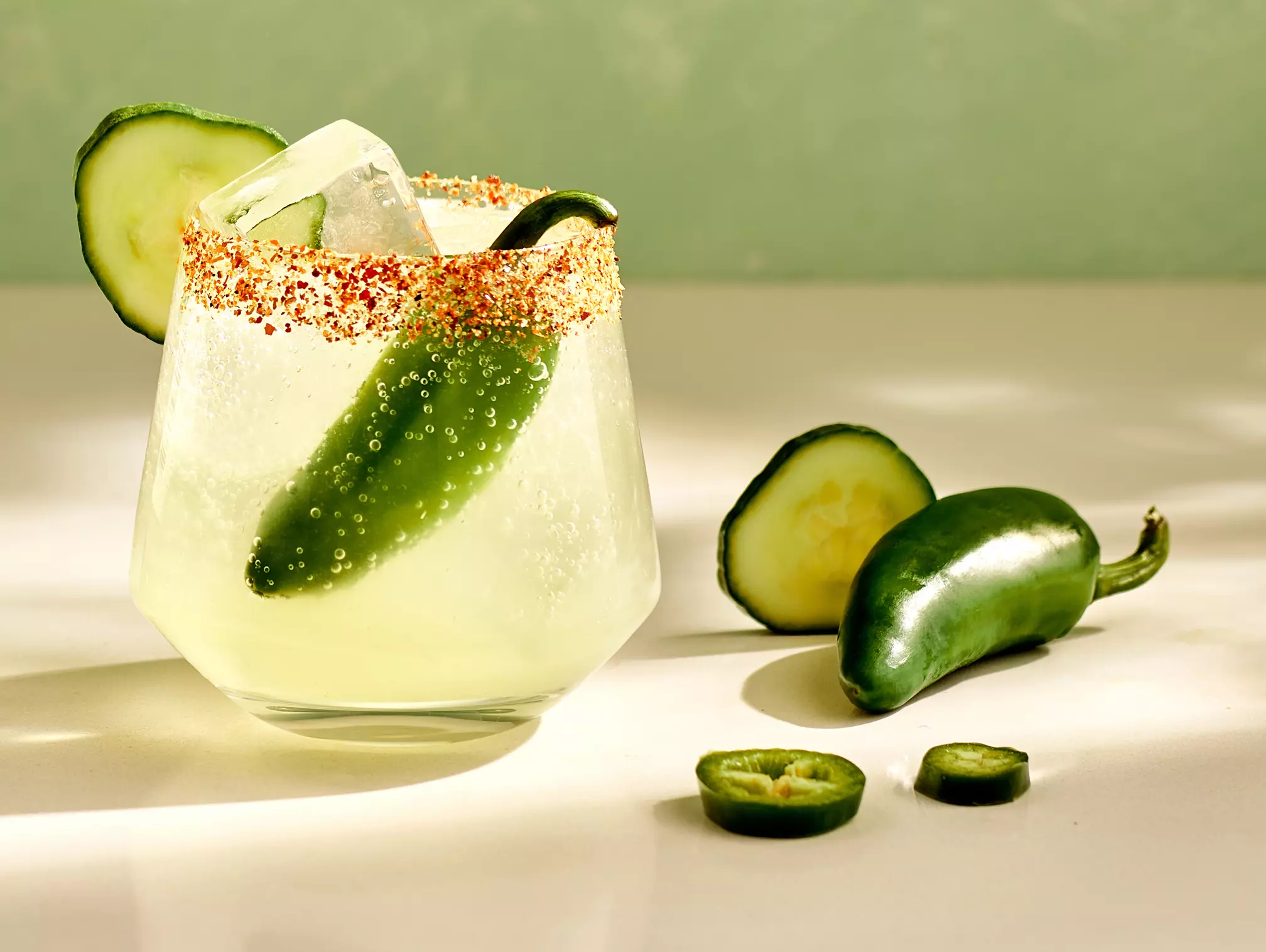 A cocktail in a glass rimmed with chili powder, garnished with a cucumber slice and a whole jalapeño, sits on a white surface next to cucumber and jalapeño slices.
