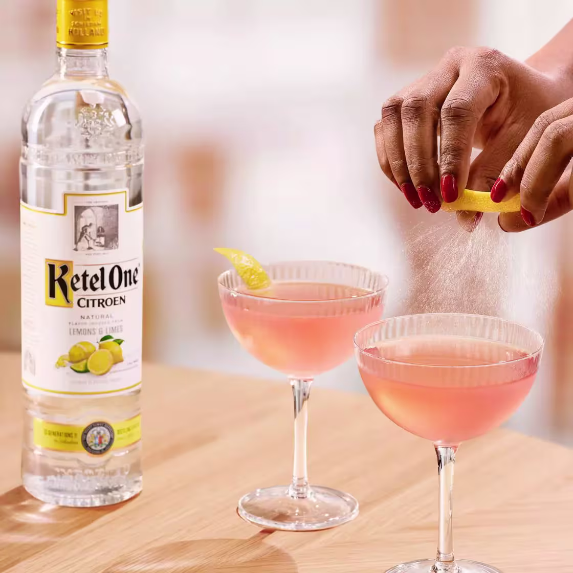 A person garnishes a pink cocktail with a lemon twist next to a bottle of Ketel One Citroen vodka on a wooden surface.