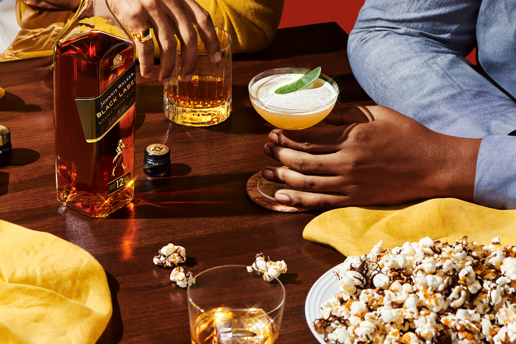 Two people sit at a table with a bottle of Johnnie Walker Black Label, two mixed drinks, a glass of whisky, and a plate of chocolate-drizzled popcorn.