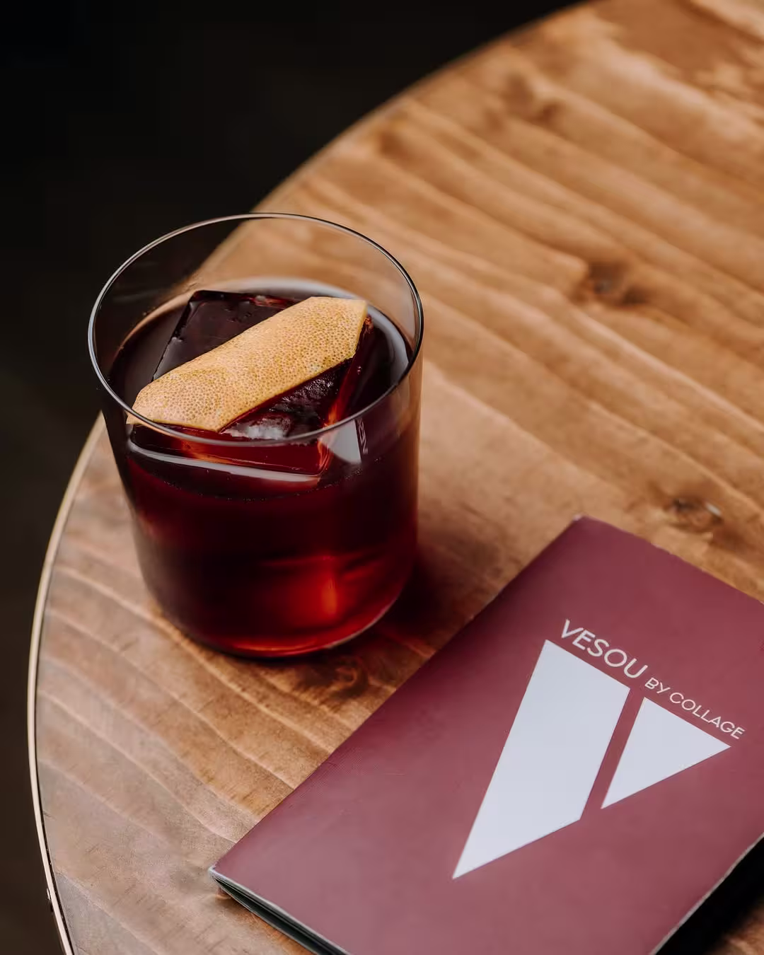 Un vaso de cóctel con hielo y una rodaja de naranja sobre una mesa de madera, junto a un menú de la coctelería Vesou by Collage, en un ambiente relajado.
